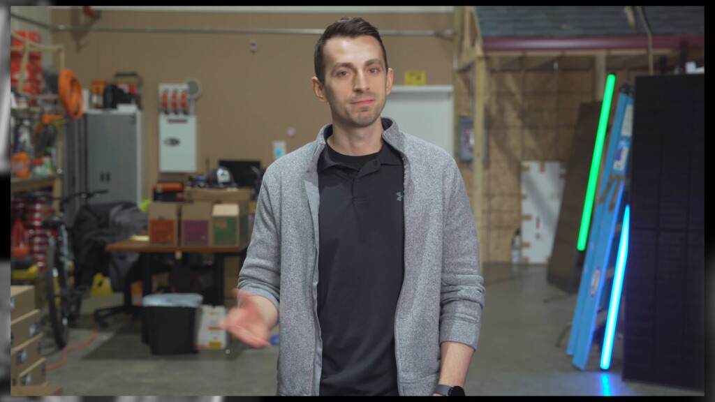 Image of a man wearing a gray cardigan and black shirt, standing in a workshop or warehouse, speaking to the camera.